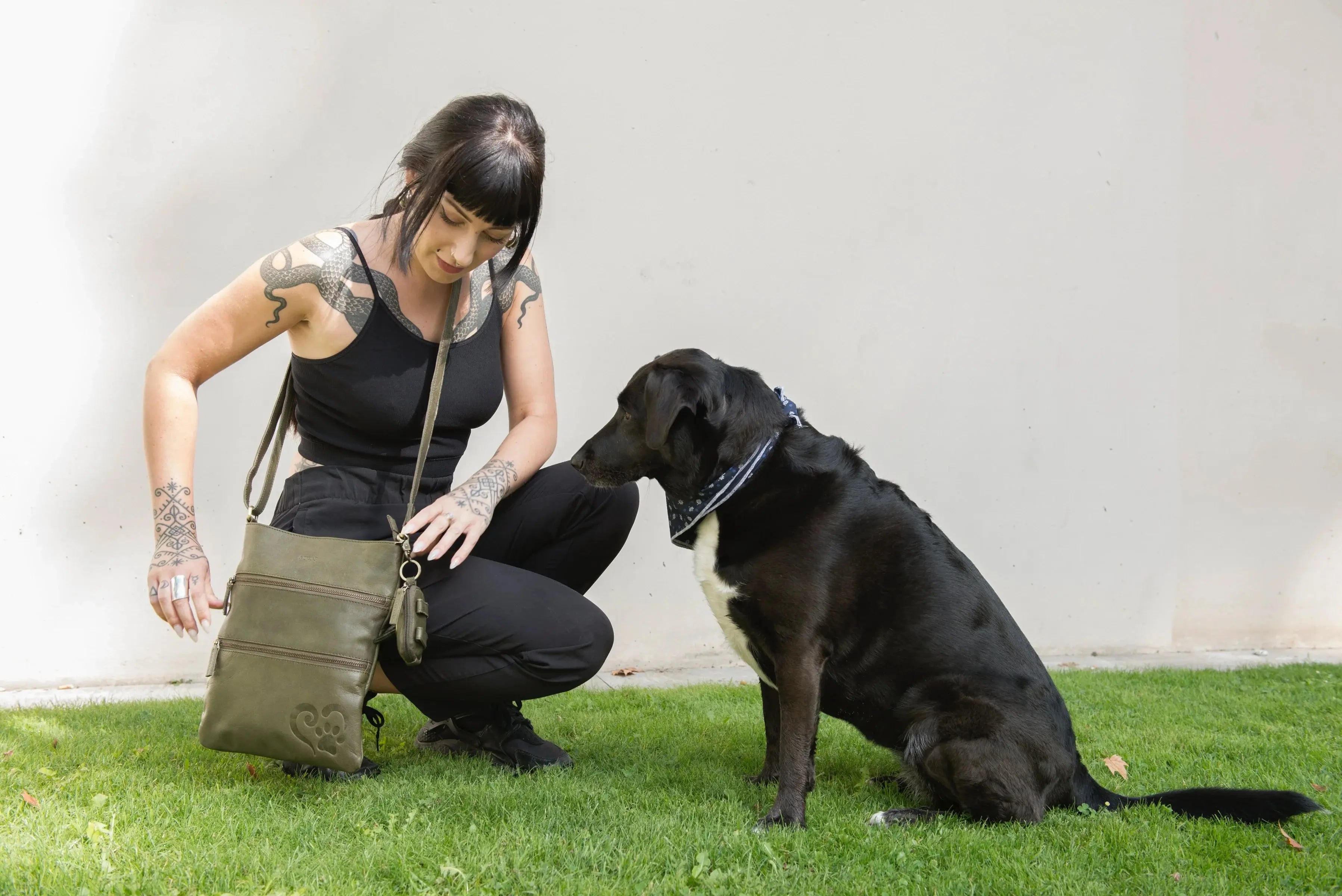 Eine Frau prĂ€sentiert hockend auf einer Wiese die Gassitasche Lisbon aus Leder in OlivgrĂŒn. Neben ihr sitzt ein Hund.
