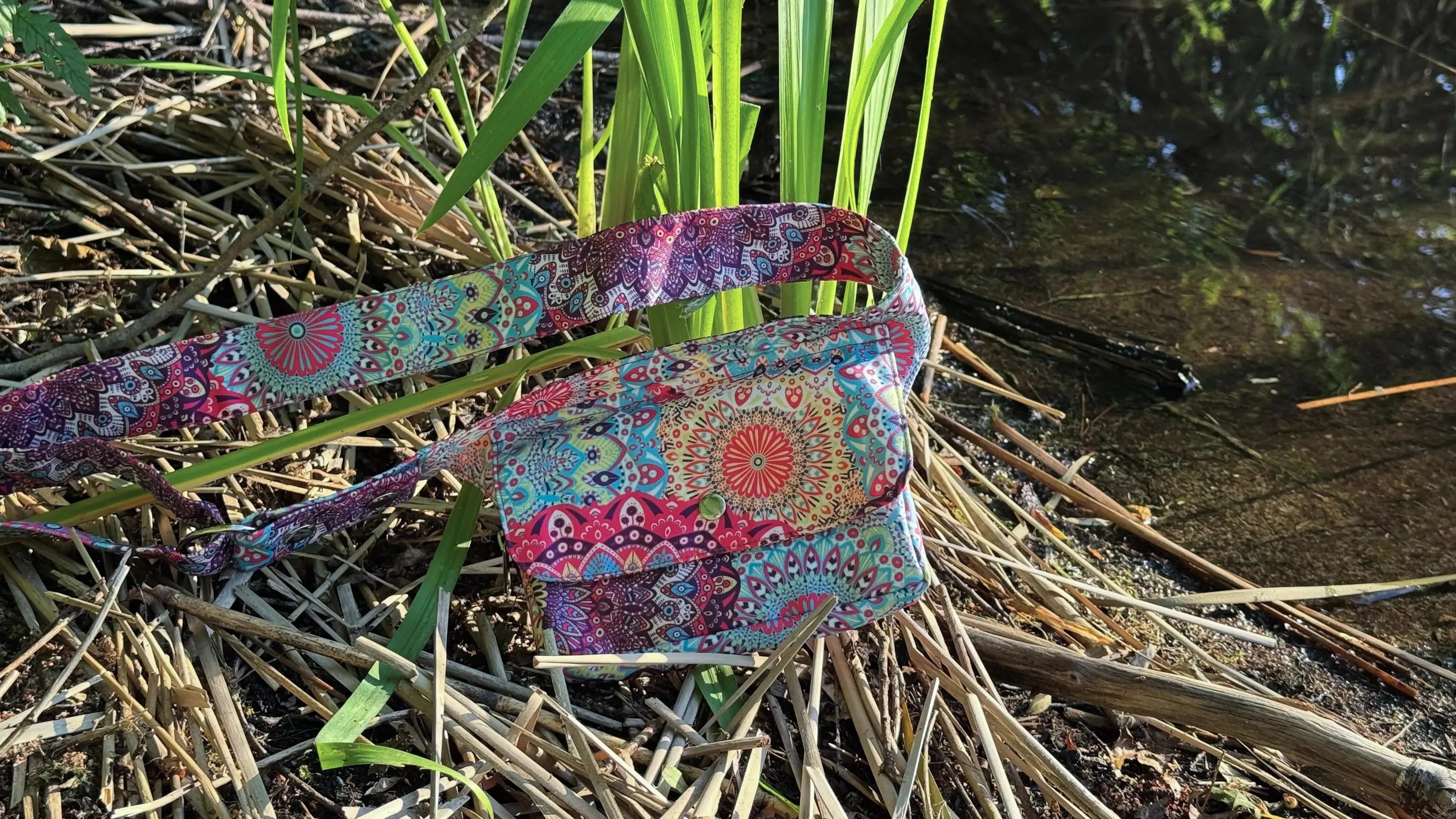 Bunte Festival-Bauchtasche mit Mandala-Muster von Nijens liegt am Ufer inmitten von Schilf und Natur, stimmungsvoll fotografiert bei Sonnenlicht.
