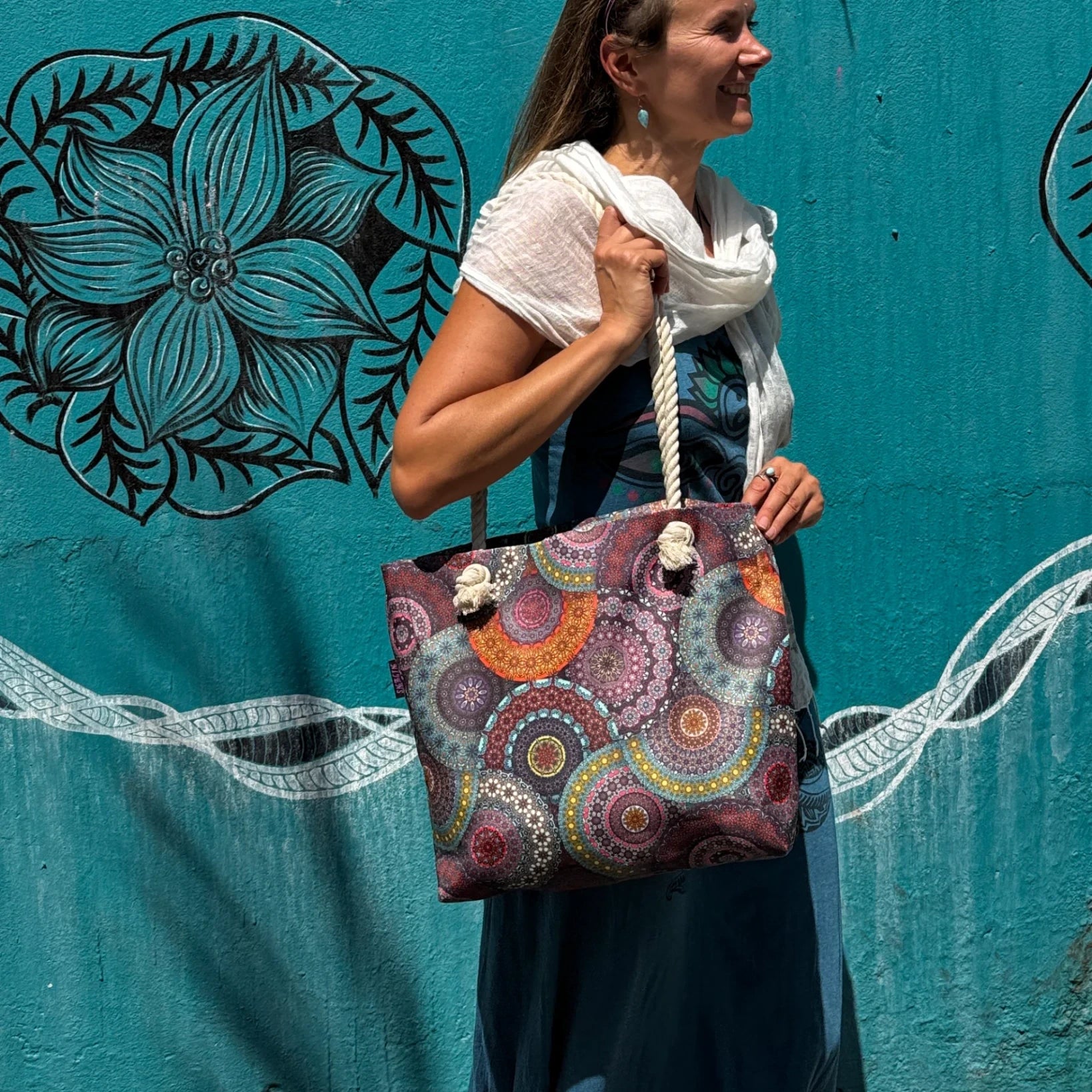 Frau mit farbenfroher Nijens Strandtasche im Mandala-Design vor türkisfarbener Wand mit Blumendekor. Bild 1:1
