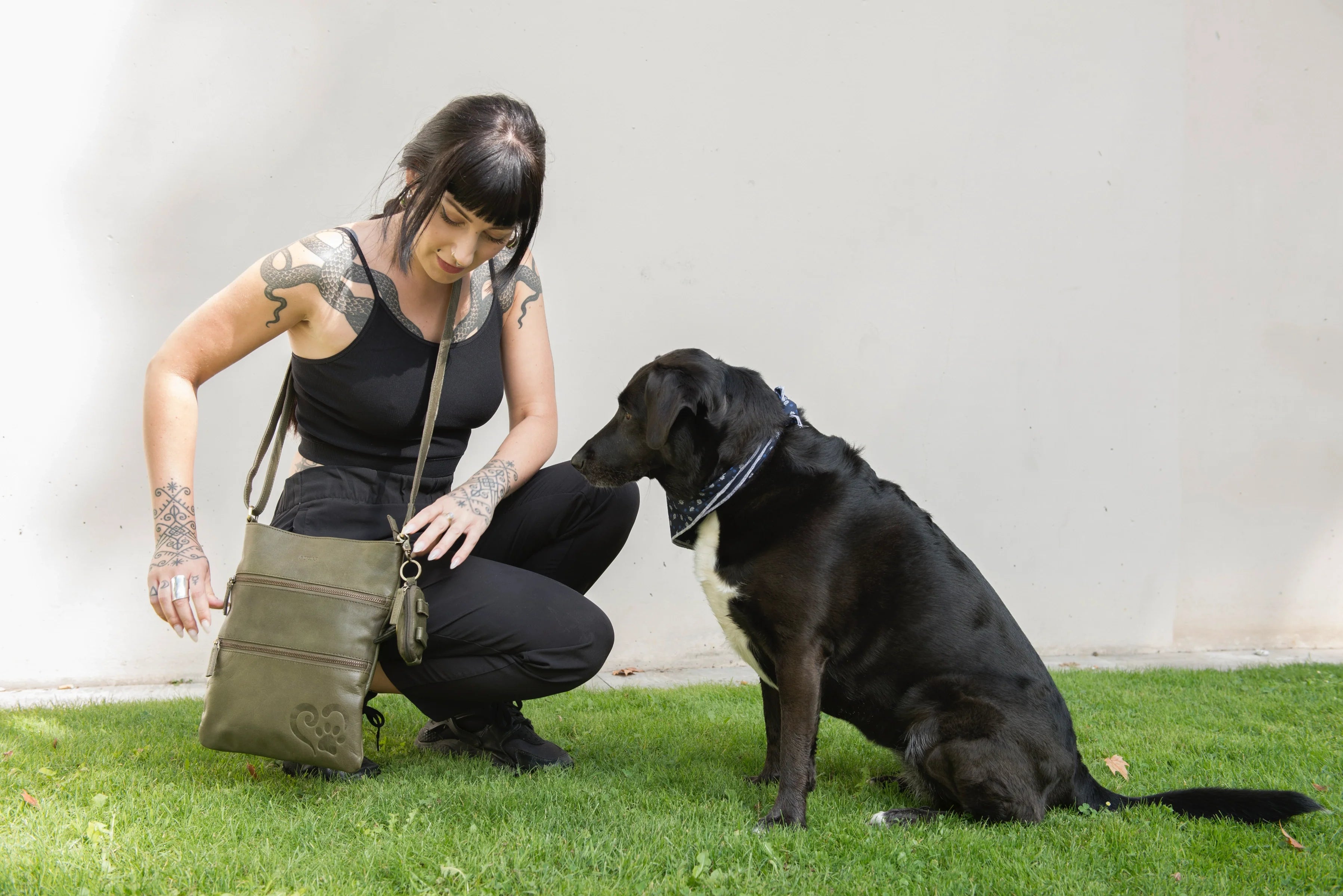 Eine Frau präsentiert hockend auf einer Wiese die Gassitasche Lisbon aus Leder in Olivgrün. Neben ihr sitzt ein Hund.