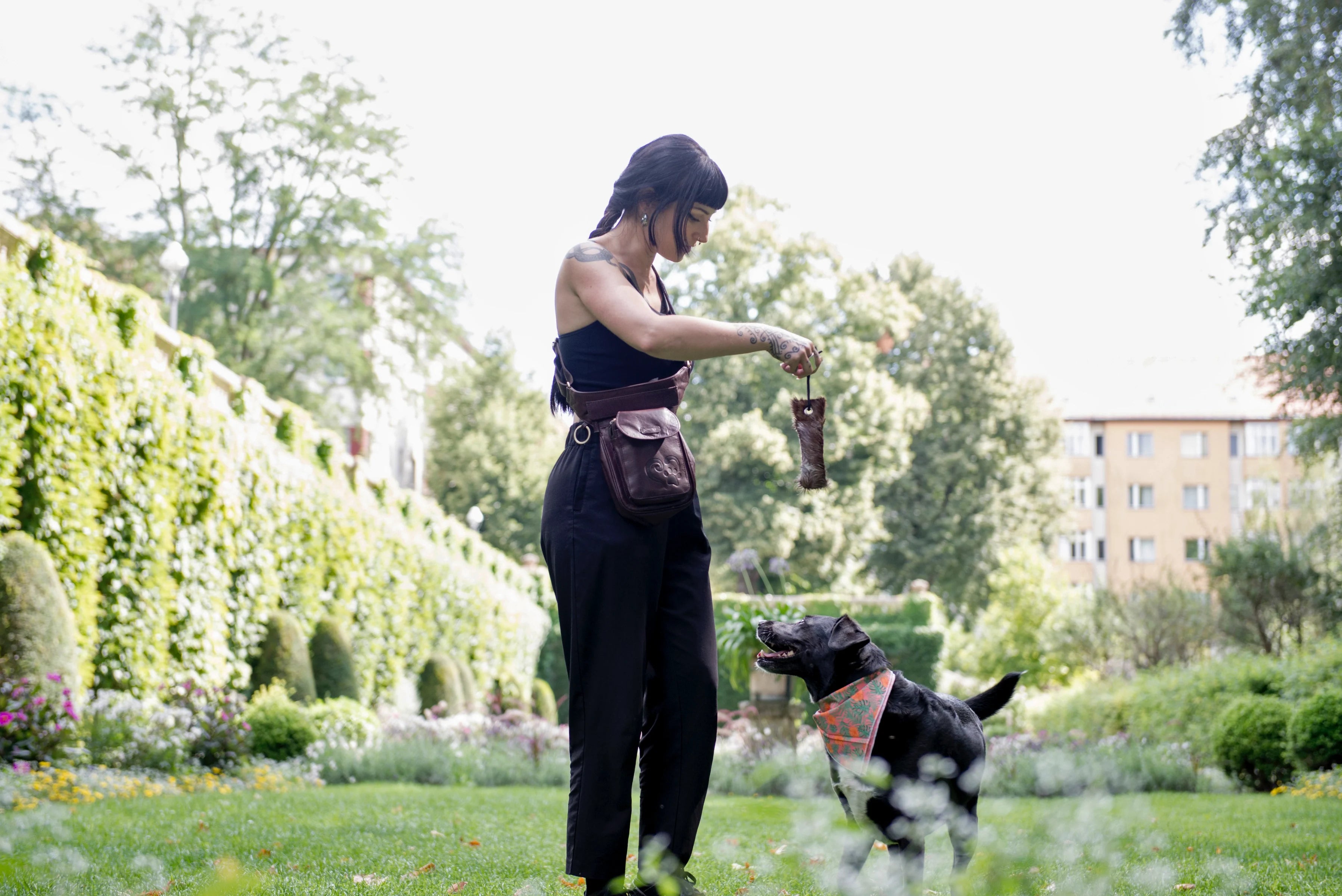 Eine Frau präsentiert spielend mit einem Hund im Park Nijens Wurfdummy DOG BONE und unsere Gassitasche Hannover in der Tragevariante Crossbody.