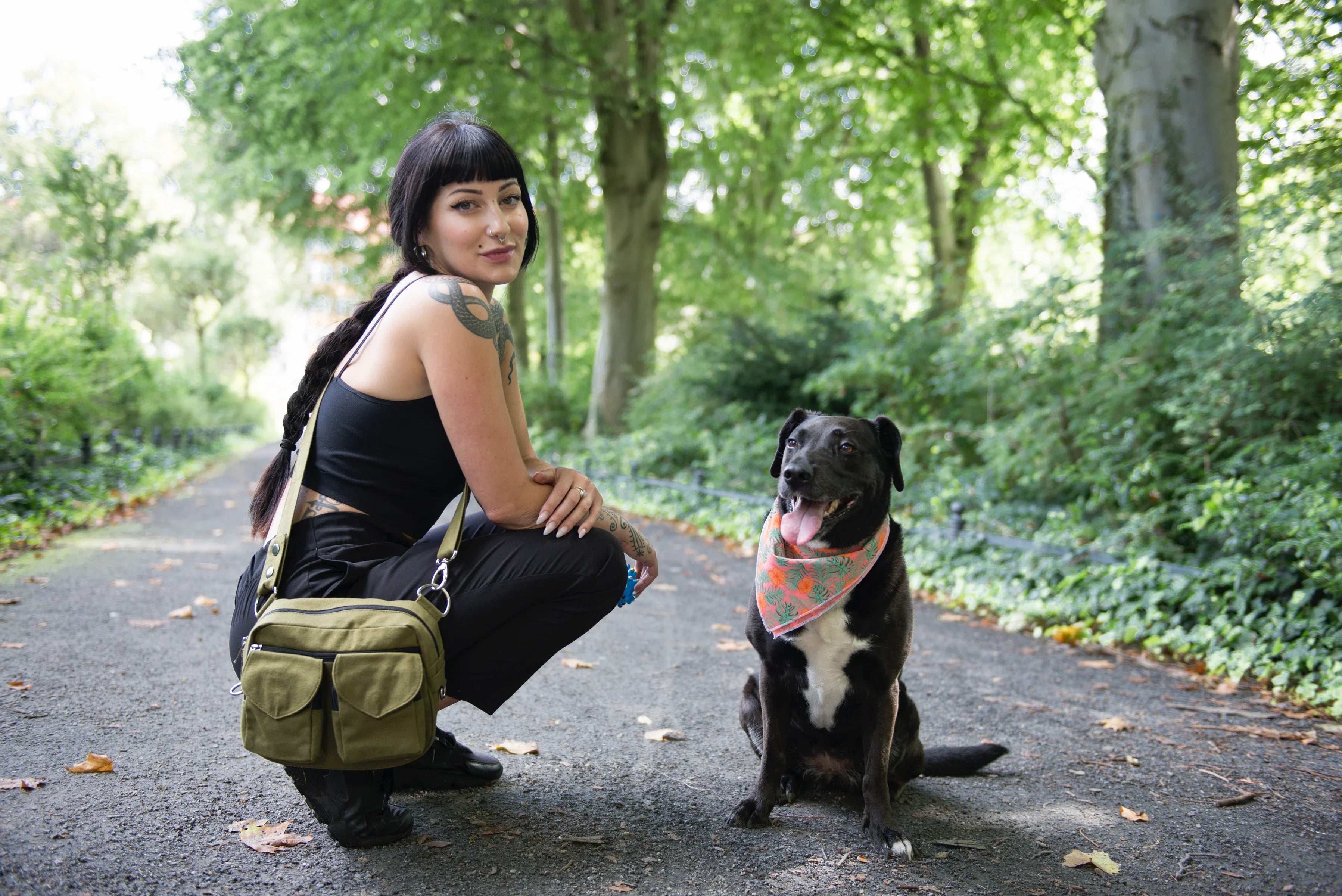 Eine Frau präsentiert hockend auf einem Parkweg unsere Gassitasche Berlin in Olivgrün. Neben ihr sitzt ein Hund mit buntem Bandana um den Hals.