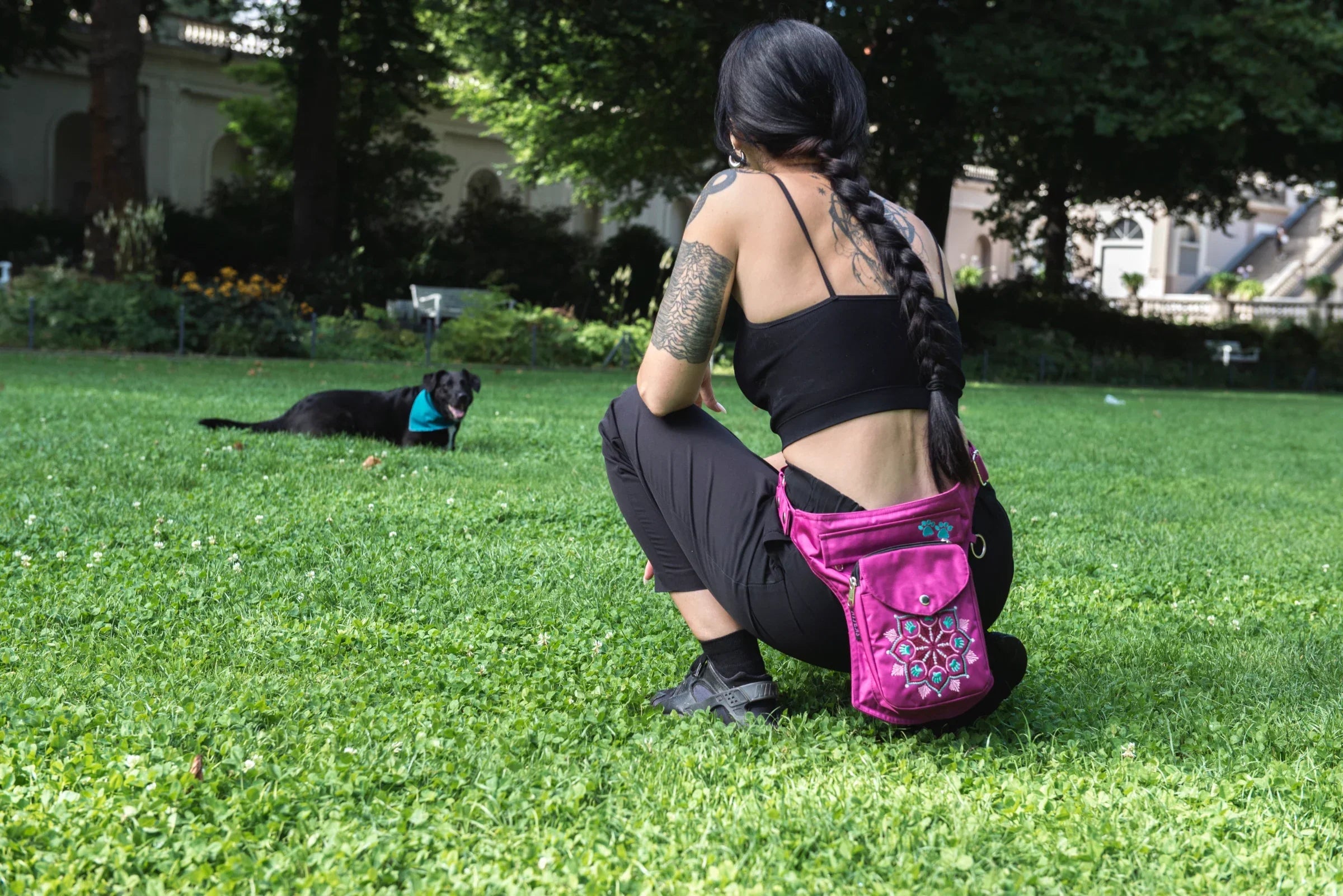 Eine Frau von hinten spielt hockend auf einer Parkwiese mit einem Hund. Um die Hüfte trägt sie Nijens Gassi-Tasche Hannover in Pink mit Mandala-Stickerei.