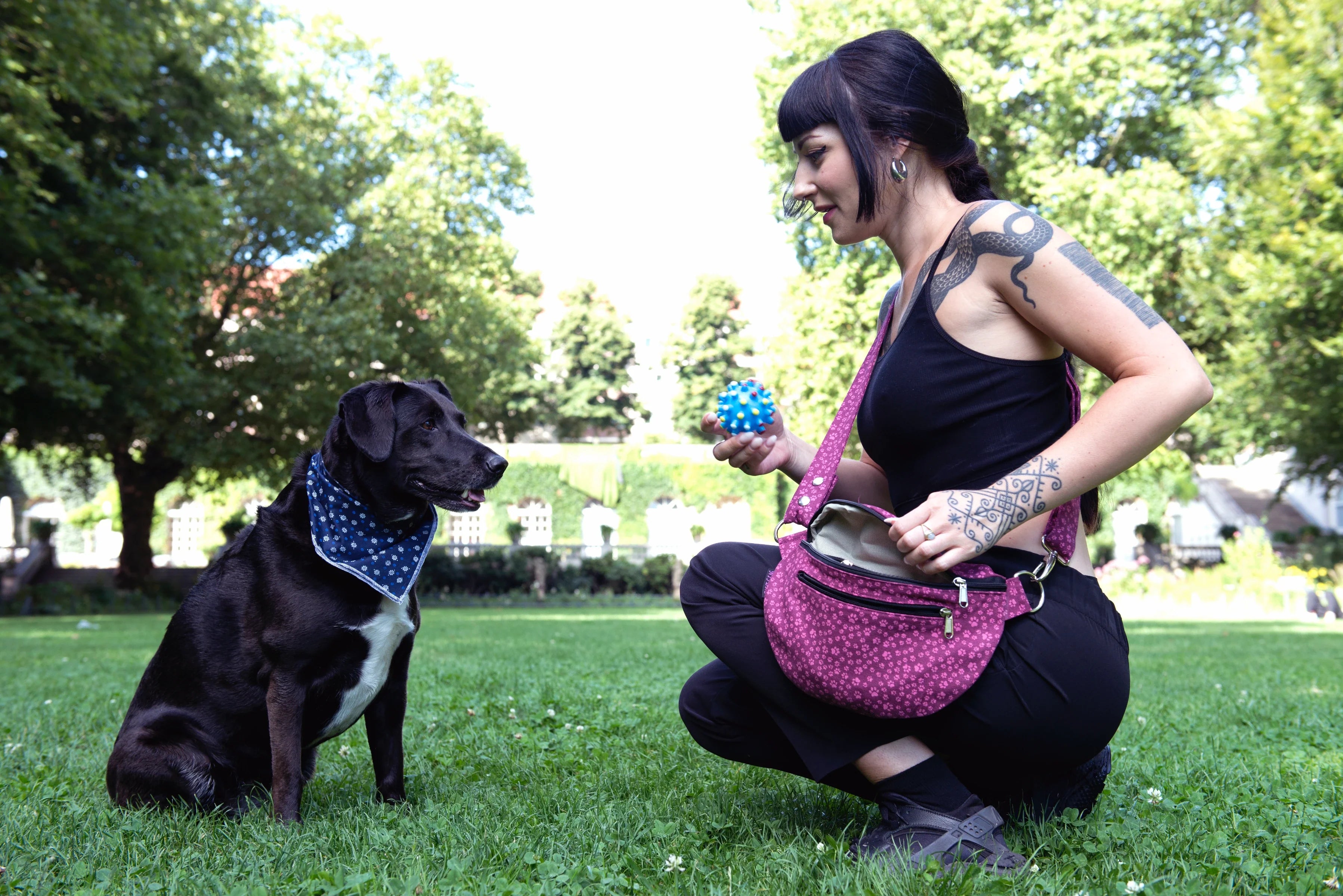 Eine Frau präsentiert hockend auf einer Parkwiese die Crossbody-Gassitasche Bangkok aus Baumwoll-Canvas in Magenta, bedruckt mit vielen kleinen Pfoten. Neben ihr sitzt ein Hund mit einem blauen Bandana um den Hals.