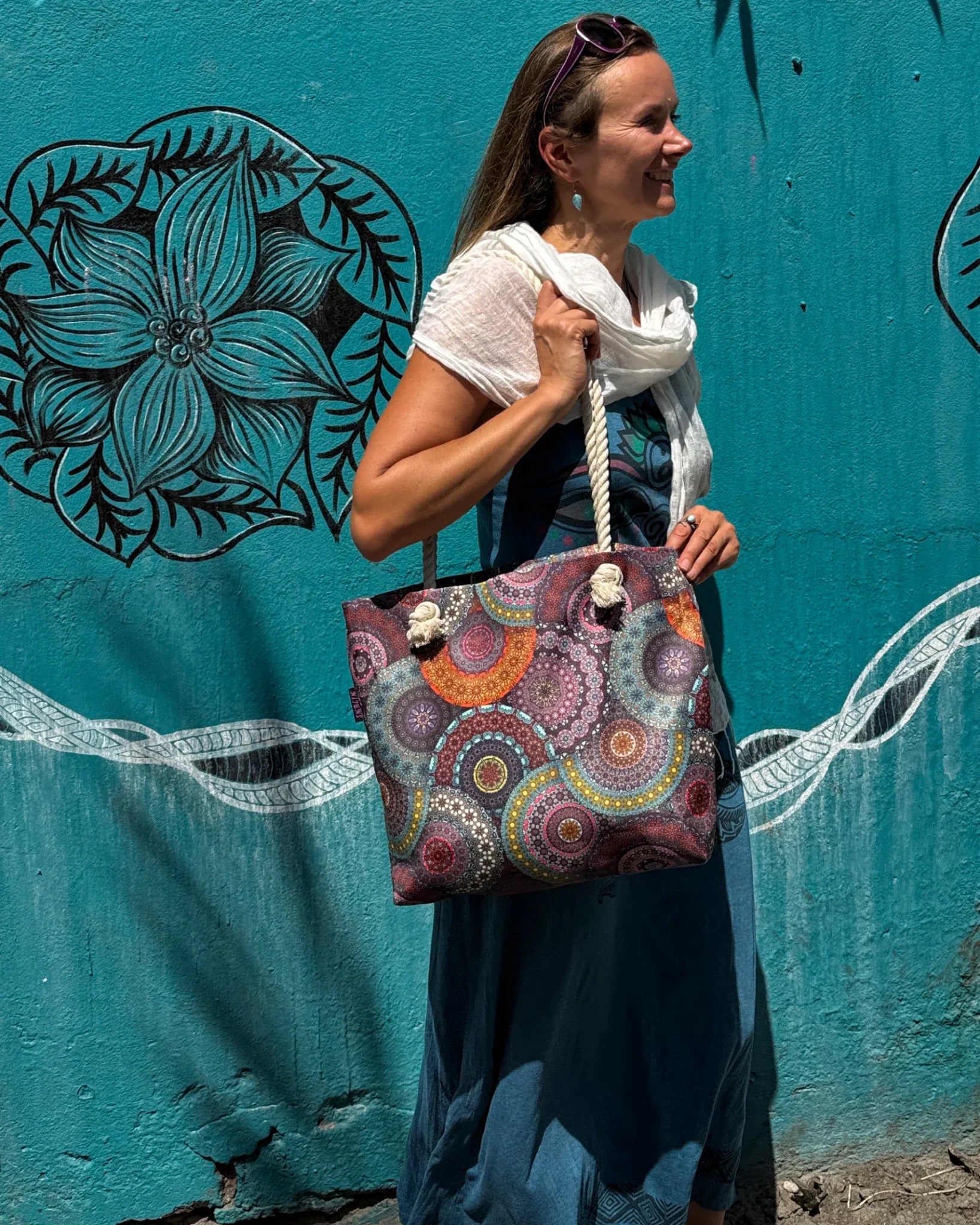 Frau mit farbenfroher Nijens Strandtasche im Mandala-Design vor türkisfarbener Wand mit Blumendekor.