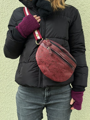 Nahaufnahme: Model präsentiert Crossbody-Bag sowie Bauchtasche aus Leder in Weinrot mit floraler Prägung. Hier wird die Gassi-Tasche als Crossbodybag über der Schulter getragen. An den Händen trägt sie Handstulpen HADAR 06 aus Schurwolle Violett.