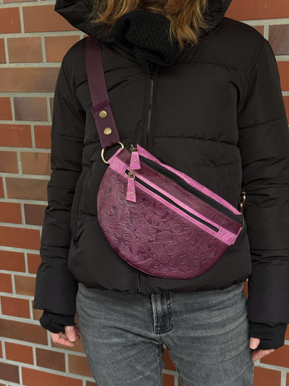 Nahaufnahme: Model präsentiert Crossbody-Bag sowie Bauchtasche aus Leder in Violett mit floraler Prägung. Hier wird die Gassi-Tasche als Crossbodybag über der Schulter getragen.