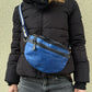 Nahaufnahme: Model präsentiert Crossbody-Bag sowie Bauchtasche aus Leder in Blau mit floraler Prägung. Hier wird die Gassi-Tasche als Crossbodybag über der Schulter getragen.