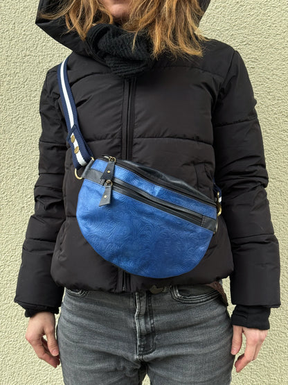 Nahaufnahme: Model präsentiert Crossbody-Bag sowie Bauchtasche aus Leder in Blau mit floraler Prägung. Hier wird die Gassi-Tasche als Crossbodybag über der Schulter getragen.