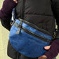 Nahaufnahme: Model präsentiert Crossbody-Bag sowie Bauchtasche aus Leder in Blau mit floraler Prägung. Hier wird die Gassi-Tasche als Crossbodybag über der Schulter getragen. An den Händen trägt sie Handstulpen HADAR 06 aus Schurwolle Violett.