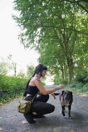 Eine Frau in schwarzer Kleidung mit  der grünen Gassi-Tasche von Nijens spielt mit einem schwarzen Hund in einem Park. 