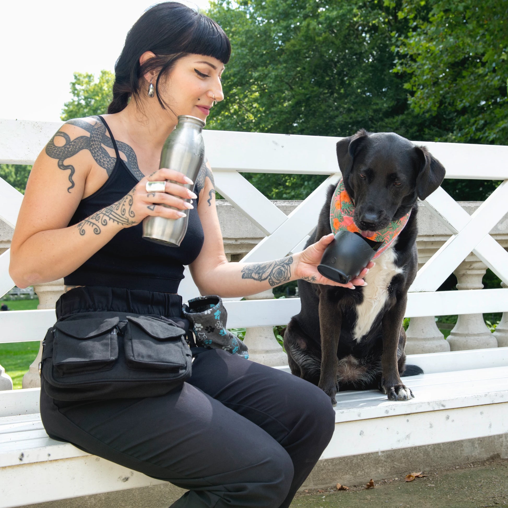 Eine Frau in schwarzer Kleidung mit  einer schwarzen Gassi-Tasche gibt einem schwarzen Hund Wasser. 