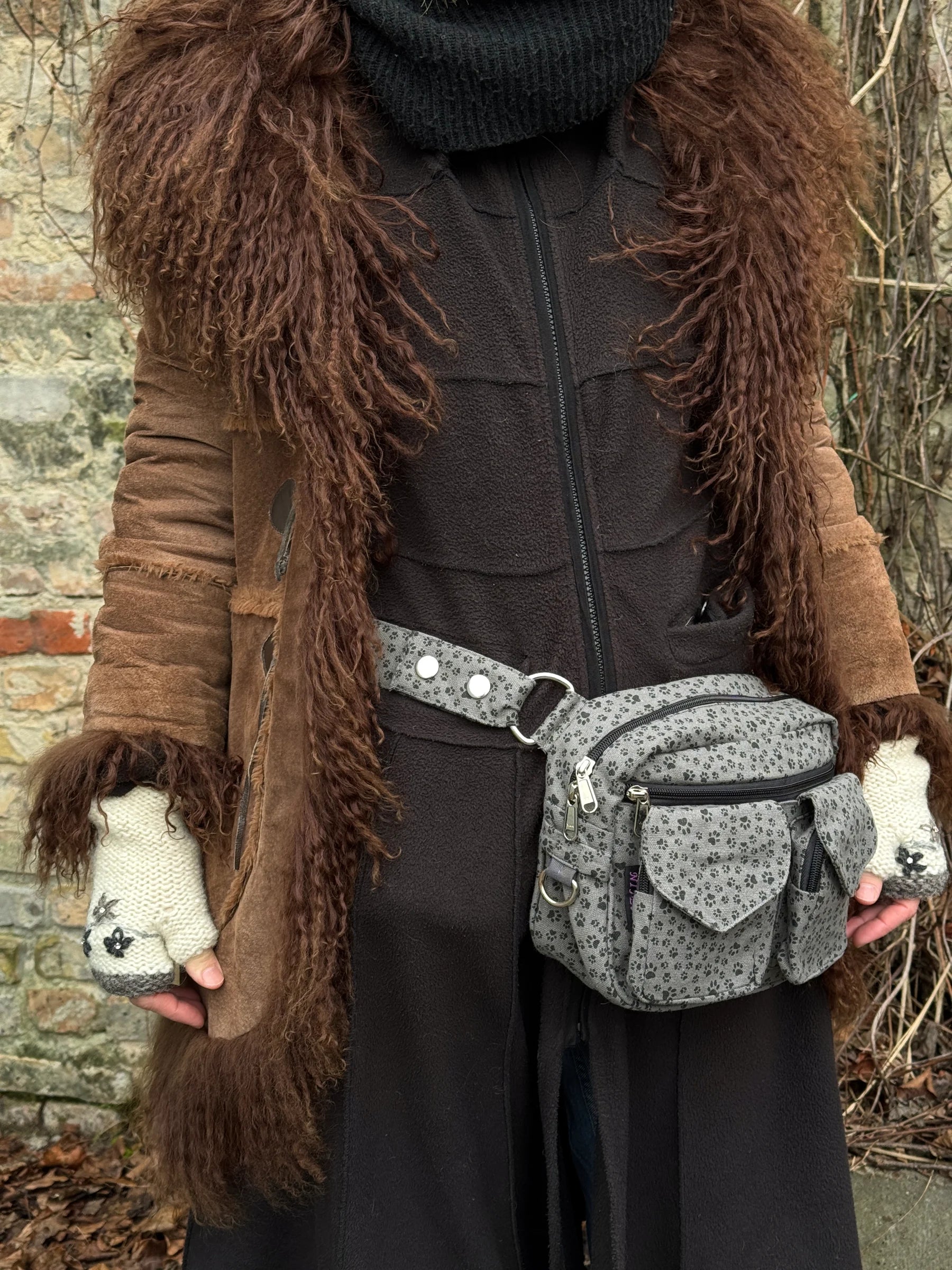 Model präsentiert Gassi-Tasche, Bauchtasche aus Baumwoll-Canvas, grau mit kleinem schwarzen Pfoten-Print. Tragevariante: Hüfttasche. An den Händen trägt sie weiße Handstulpen mit Blumenstickerei und Strass aus Schurwolle.