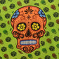 Grüne Tagesdecke bedruckt mit großen orangen mexikanischen Totenkopf in der Mitte. Totenkopf hat jeweils eine Blumenbemalung mit Blumen auf dem Kopf und in den Augen.