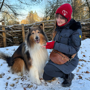 Frau im Winterpark mit Collie-Hund trägt rote Schurwollmütze mit Pfotenmotiv, passende Armstulpen und braune Leder-Gürteltasche von Nijens.