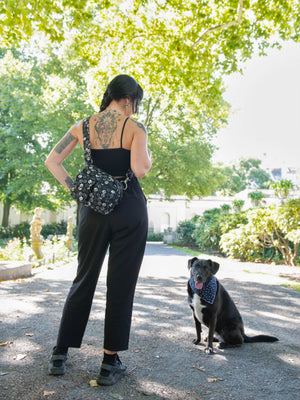Eine Frau in schwarzer Kleidung mit  Gassi-Tasche spielt mit einem schwarzen Hund in einem Park. Nijens UmhĂ€ngetasche fĂŒr den Ausflug mit Hund.