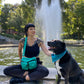 Eine Frau in schwarzer Kleidung mit einer türkisfarbenen Gassi-Tasche spielt mit einem schwarzen Hund vor einem Springbrunnen in einem Park.