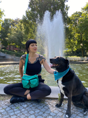 Eine Frau in schwarzer Kleidung mit einer türkisfarbenen Gassi-Tasche spielt mit einem schwarzen Hund vor einem Springbrunnen in einem Park.