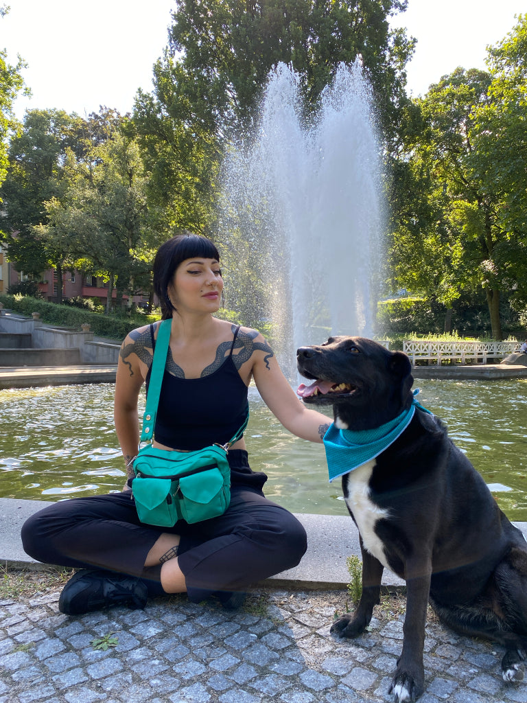 Eine Frau in schwarzer Kleidung mit einer türkisfarbenen Gassi-Tasche spielt mit einem schwarzen Hund vor einem Springbrunnen in einem Park.