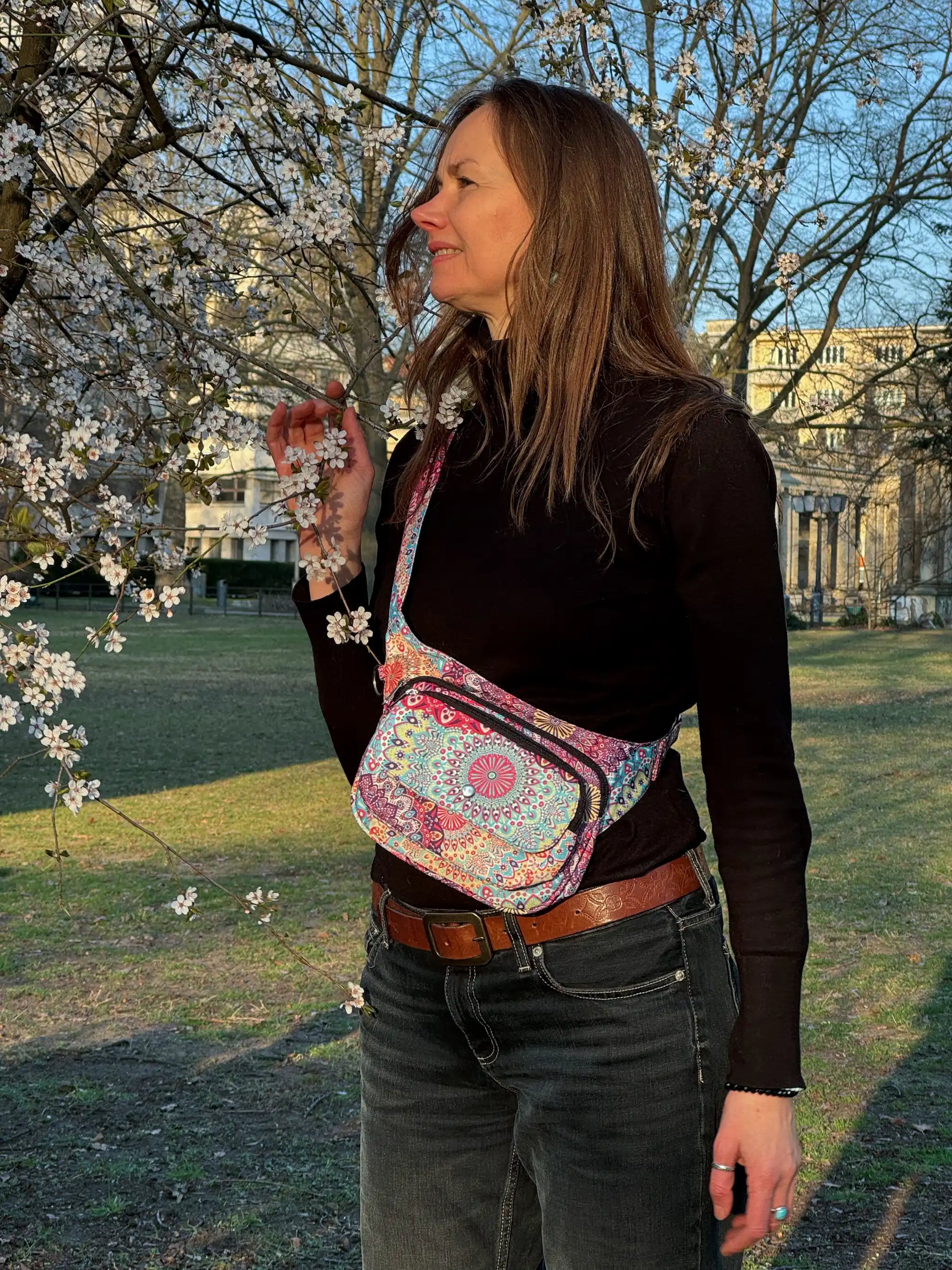 Frau trägt die bunte Mandala-Hüfttasche von Nijens als Crossbody-Bag über der Schulter, im Park mit blühenden Bäumen.

