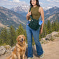 Frau mit tannengrüner Gassi-Umhängetasche und goldenem Retriever auf einem Bergweg vor alpiner Kulisse mit Bergen.