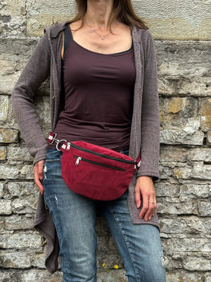 Frau trägt eine weinrote Crossbody Bag aus Cord mit Reißverschlussfächern lässig vor einer Natursteinmauer.