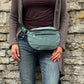 Frau trägt eine salbeigrüne Crossbody Bag aus Cord mit Reißverschlussfächern lässig vor einer Natursteinmauer.