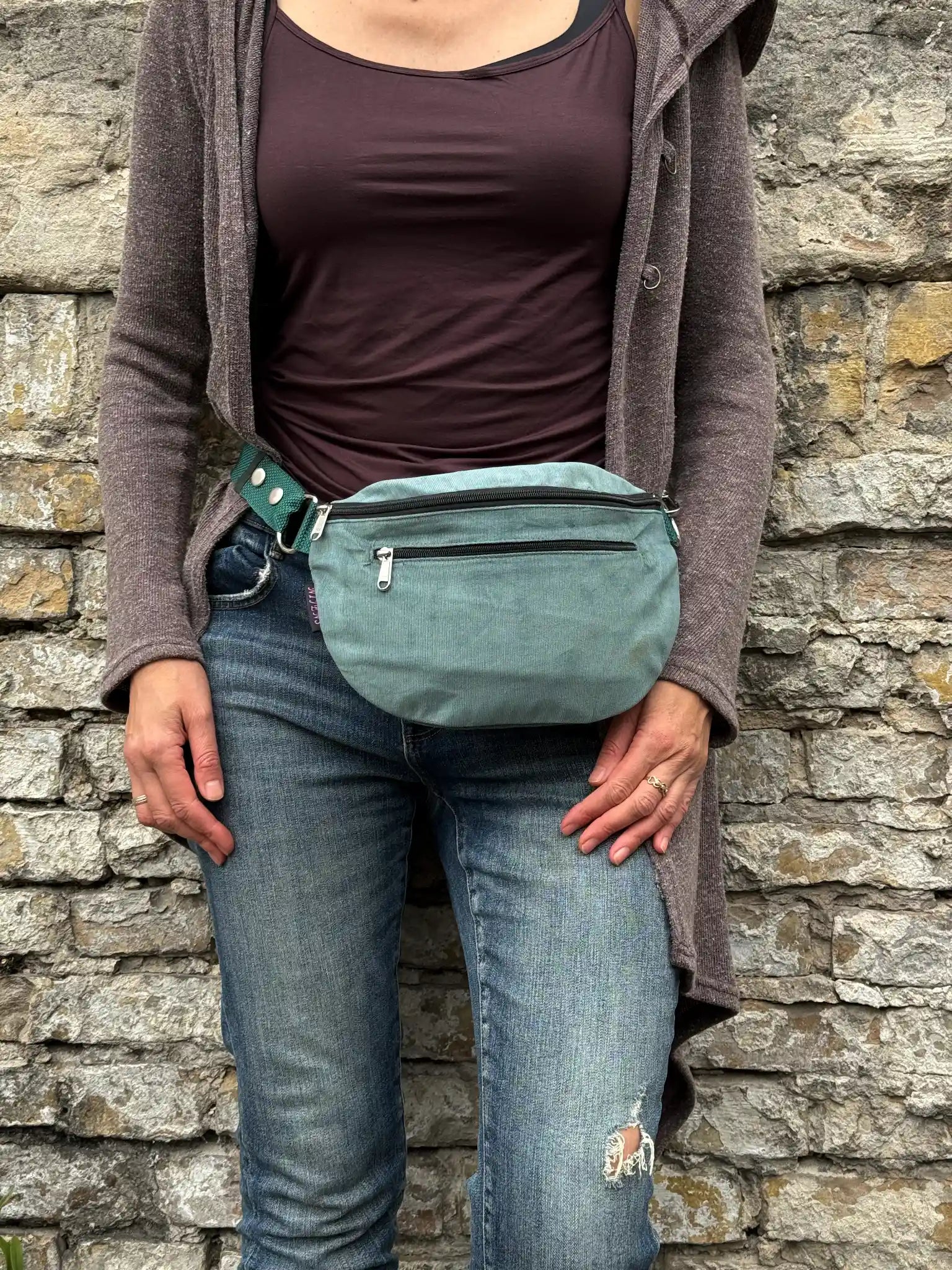 Frau trägt eine salbeigrüne Crossbody Bag aus Cord mit Reißverschlussfächern lässig vor einer Natursteinmauer.