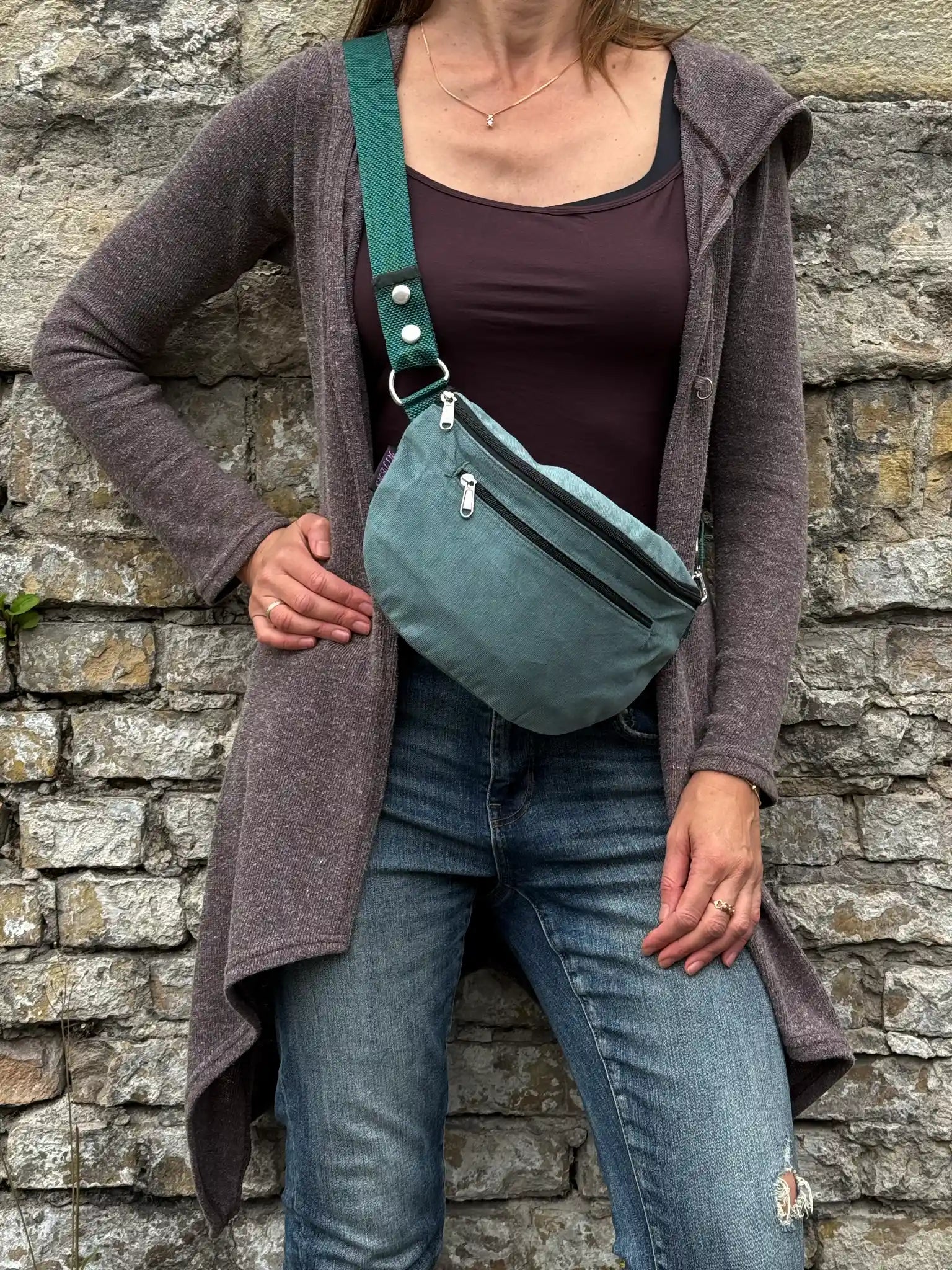 Frau trägt salbeigrüne Cord Crossbody Bag lässig über der Schulter vor einer Natursteinmauer