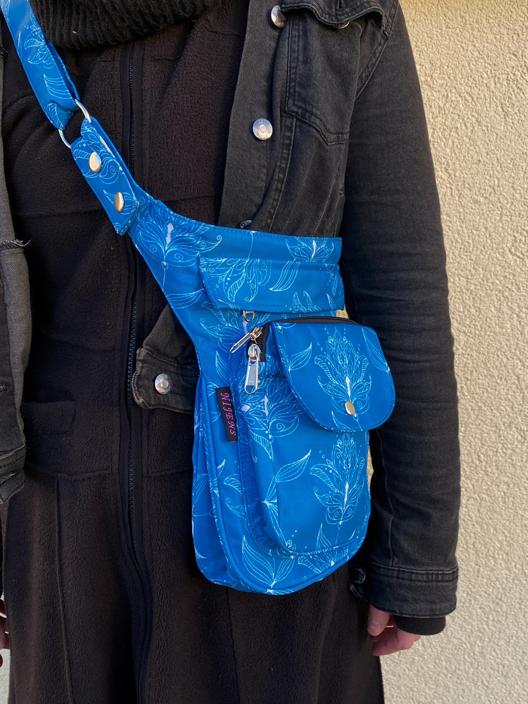 Model präsentiert Gassi-Tasche, Bauchtasche und Gürteltasche aus Polyesterblau mit floralen Elementen in der Tragevariante Crossbodybag.