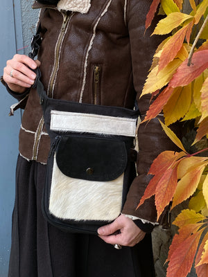 Nahansicht: Model vor herbstlicher Kulisse mit Gürteltasche, Gassi-Tasche in Canvas-Schwarz, Reißverschlussklappe und Vordertasche sind mit weißem Kuhfell und Leder schwarz besetzt. Diese Gürteltasche hat zwei Reißverschlussfächer vorne sowie ein Einsteckfach, welches durch einen Überschlag mit Druckknopf verschlossen wird. Auf der hinteren Seite befindet sich ein weiteres Reißverschlussfach. An der Seite des Taschengurtes befindet sich ein kleiner Metallring zum Anhängen kleiner Beuteltaschen. 