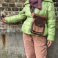 Ausschnitt: Model präsentiert die Hüfttasche, Gassi-Tasche aus Leder und Baumwoll-Canvas in Brauntönen, mit floraler Ornamentenprägung in der Tragevariante Crossbody-Bag. Diese Hüfttasche hat ein Reißverschlussfach auf der hinteren Seite und zwei Reißverschlussfächer vorne sowie ein Einsteckfach, welches durch einen Überschlag mit einem Magnetknopf verschlossen wird. An der Seite des Taschengurtes befindet sich ein kleiner Metallring in Form eines Dreiecks zum Anhängen kleiner Beuteltaschen.