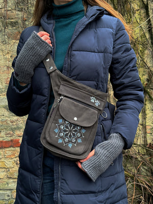 Ein Model präsentiert eine Gürteltasche aus Canvas-Stoff in Dunkelgrau mit Pfoten-Mandala-Stickerei. An den Händen trägt sie Handstulpen MAHALAXMI 54 aus Schurwolle in Grau.