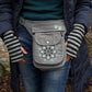 Nahaufnahme: Model präsentiert Gassi-Tasche, Bauchtasche und Gürteltasche aus Canvas-Stoff grau mit Pfoten-Mandala-Stickerei. An den Händen trägt sie gestreifte Handstulpen HADAR C-27 aus Schurwolle hellgrau, schwarz, mit Daumenspalten.