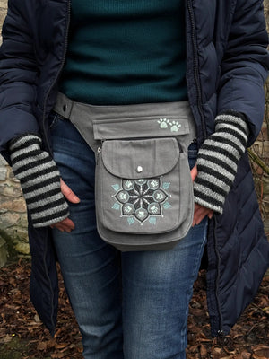 Nahaufnahme: Model präsentiert Gassi-Tasche, Bauchtasche und Gürteltasche aus Canvas-Stoff grau mit Pfoten-Mandala-Stickerei. An den Händen trägt sie gestreifte Handstulpen HADAR C-27 aus Schurwolle hellgrau, schwarz, mit Daumenspalten.