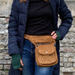Model präsentiert Bauchtasche, Gassi-Tasche aus Cord Goldbraun. Diese Gürteltasche hat zwei Reißverschlussfächer vorne sowie ein Einsteckfach, welches durch einen Überschlag mit Druckknopf verschlossen wird. Auf der hinteren Seite befindet sich ein weiteres Reißverschlussfach. An der Seite des Taschengurtes befindet sich ein kleiner Metallring, zum anhängen kleiner Beuteltaschen.