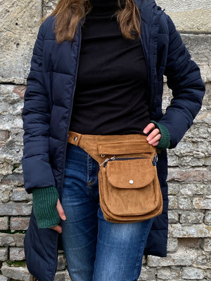 Model präsentiert Bauchtasche, Gassi-Tasche aus Cord Goldbraun. Diese Gürteltasche hat zwei Reißverschlussfächer vorne sowie ein Einsteckfach, welches durch einen Überschlag mit Druckknopf verschlossen wird. Auf der hinteren Seite befindet sich ein weiteres Reißverschlussfach. An der Seite des Taschengurtes befindet sich ein kleiner Metallring, zum anhängen kleiner Beuteltaschen.