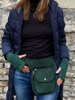 Model präsentiert Bauchtasche, Gassi-Tasche aus Cord Tannengrün. Diese Gürteltasche hat zwei Reißverschlussfächer vorne sowie ein Einsteckfach, welches durch einen Überschlag mit Druckknopf verschlossen wird. Auf der hinteren Seite befindet sich ein weiteres Reißverschlussfach. An der Seite des Taschengurtes befindet sich ein kleiner Metallring, zum anhängen kleiner Beuteltaschen. An den Händen trägt das Model MAHALAXMI 53 Handstulpen mit Daumenspalte aus Schurwolle Tannengrün.