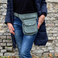 Model präsentiert Bauchtasche, Gassi-Tasche aus Cord Salbeigrün. Diese Gürteltasche hat zwei Reißverschlussfächer vorne sowie ein Einsteckfach, welches durch einen Überschlag mit Druckknopf verschlossen wird. Auf der hinteren Seite befindet sich ein weiteres Reißverschlussfach. An der Seite des Taschengurtes befindet sich ein kleiner Metallring, zum anhängen kleiner Beuteltaschen.