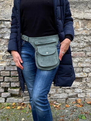 Model präsentiert Bauchtasche, Gassi-Tasche aus Cord Salbeigrün. Diese Gürteltasche hat zwei Reißverschlussfächer vorne sowie ein Einsteckfach, welches durch einen Überschlag mit Druckknopf verschlossen wird. Auf der hinteren Seite befindet sich ein weiteres Reißverschlussfach. An der Seite des Taschengurtes befindet sich ein kleiner Metallring, zum anhängen kleiner Beuteltaschen.