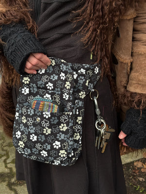 Nahaufnahme Bauchtasche: Model präsentiert Gassi-Tasche, Bauchtasche Hannover aus Baumwoll-Canvas in Schwarz mit großen Pfoten-Prints. Vorne befindet sich ein verschließbares Einsteckfach. Am Gurt gibt es einen Schlüsselring, an dem ein Schlüssel mit Karabiner hängt. Sie trägt an den Händen Handstulpen mit Daumen aus Schurwolle, schwarz.