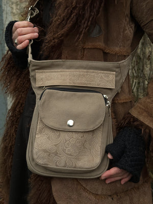 Nahaufnahme von Tasche: Model präsentiert Gürteltasche, Gassi-Tasche aus Baumwoll-Canvas, sandfarben. Reißverschlussklappe und Vordertasche sind mit Blumen geprägtem Leder in Beige besetzt. Tragevariante: Crossbodybag.