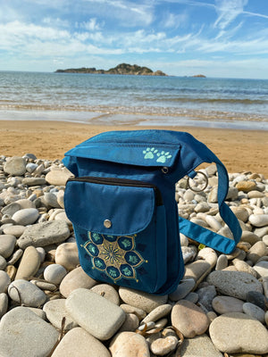 Gassitasche, Bauchtasche aus wasserabweisendem Nylonstoff Petrol-Türkis mit Blumenpfotenmandala-Stickerei. Am Ufer gegen das blaue Meer.