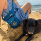Gassitasche für Hundebesitzer, Bauchtasche aus wasserabweisendem Nylonstoff Petrol-Türkis mit Blumenpfotenmandala-Stickerei. Am Ufer gegen das blaue Meer.