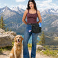 Frau mit lodengrüner Gassi-Tasche und goldenem Retriever auf einem Bergweg vor alpiner Kulisse mit See und schneebedeckten Bergen.