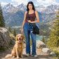 Frau mit lodengrüner Gassi-Umhängetasche und goldenem Retriever auf einem Bergweg vor alpiner Kulisse mit See und schneebedeckten Bergen.
