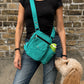 Frau mit schwarzem Oberteil und Jeans trägt eine türkisfarbene Nijens Gassi-Tasche mit Flaschenfach, während ein cremefarbener Hund zur Tasche hochschaut.