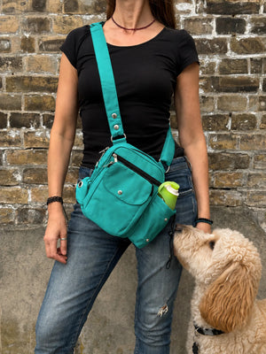 Frau mit schwarzem Oberteil und Jeans trägt eine türkisfarbene Nijens Gassi-Tasche mit Flaschenfach, während ein cremefarbener Hund zur Tasche hochschaut.