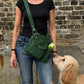 Frau mit schwarzem Oberteil und Jeans trägt eine grüne Nijens Gassi-Tasche mit Flaschenfach, während ein cremefarbener Hund zur Tasche hochschaut.
