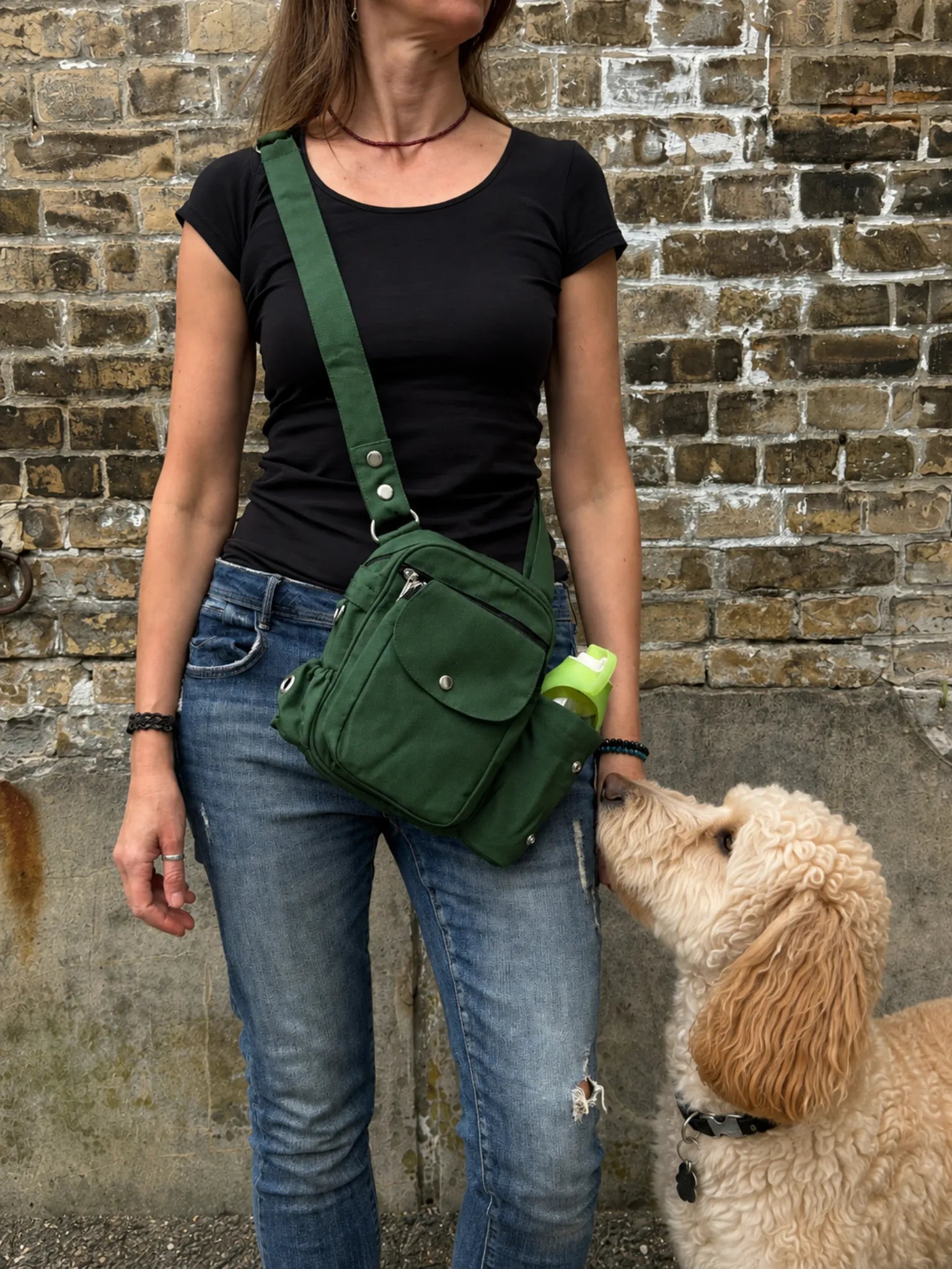 Frau mit schwarzem Oberteil und Jeans trägt eine grüne Nijens Gassi-Tasche mit Flaschenfach, während ein cremefarbener Hund zur Tasche hochschaut.